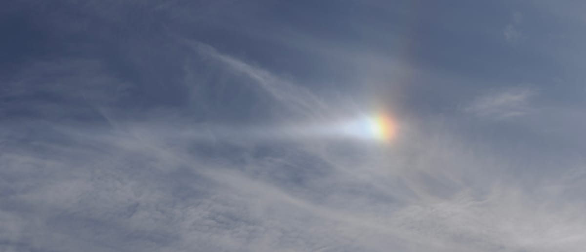 A picture of a blue, but cloudy sky. The sun is refracting through the clouds to create a rainbow blob in the centre, with a shaft of bright white to the left.